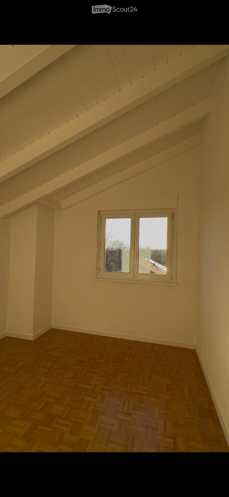 Empty attic room with wooden herringbone flooring, white walls, and a single window offering a view of the outside.