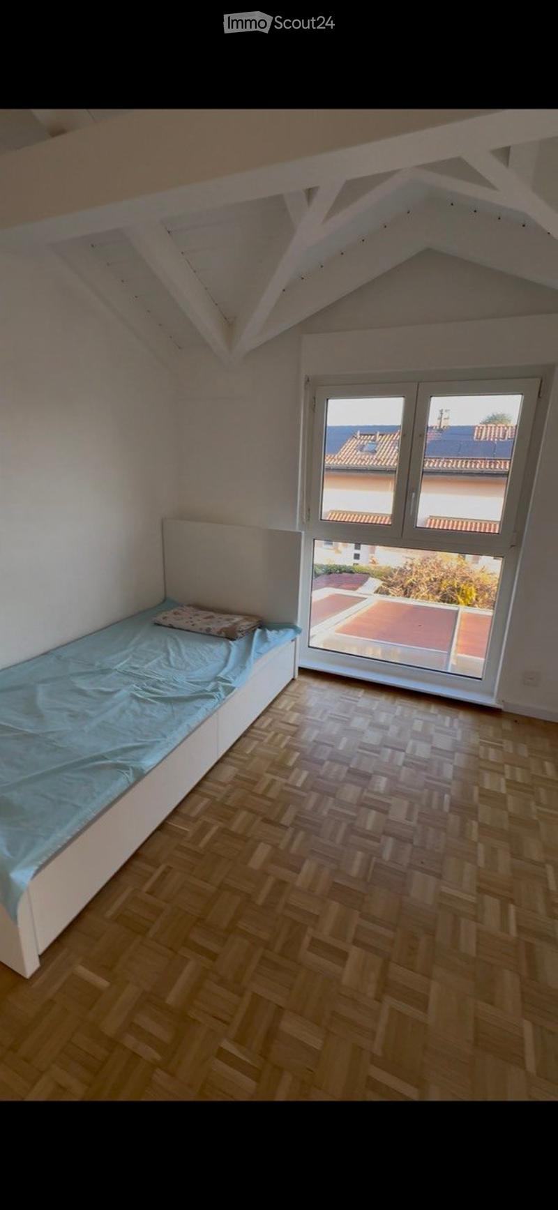 Spacious bedroom with wooden herringbone flooring, white walls, and a large window offering a view of the roof and surrounding area. The room features a single bed with a blue duvet and a pillow, positioned against the wall.