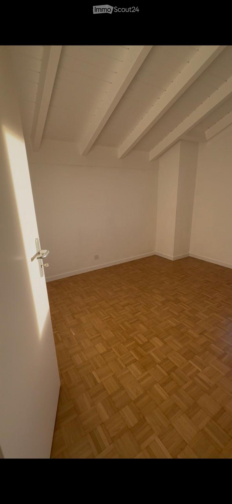 Empty room with wooden herringbone flooring, white walls, and exposed wooden beams on the ceiling. No furniture or equipment present.
