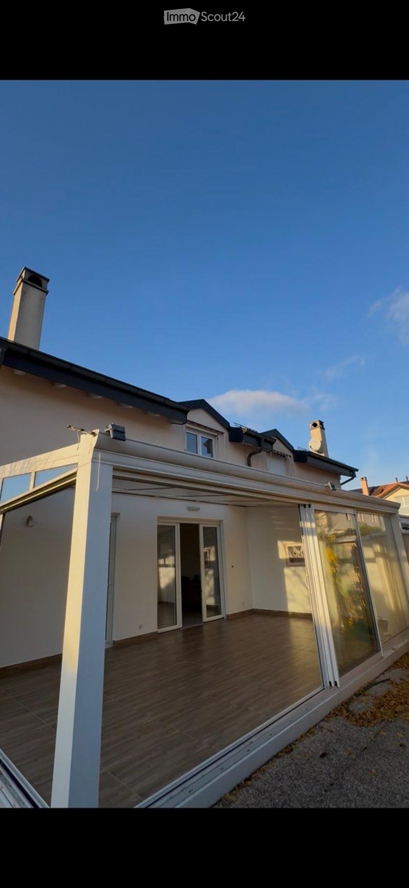 Modern wooden deck terrace with glass railings, attached to a house with multiple chimneys and windows.