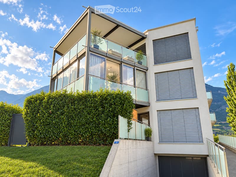 Modern two-story house with large windows, glass balcony railings, and a well-maintained garden.