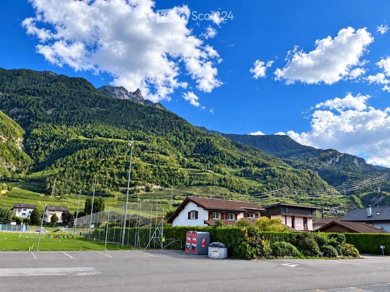 A small village with houses and a parking lot in the foreground, surrounded by lush green hills and mountains under a clear blue sky.