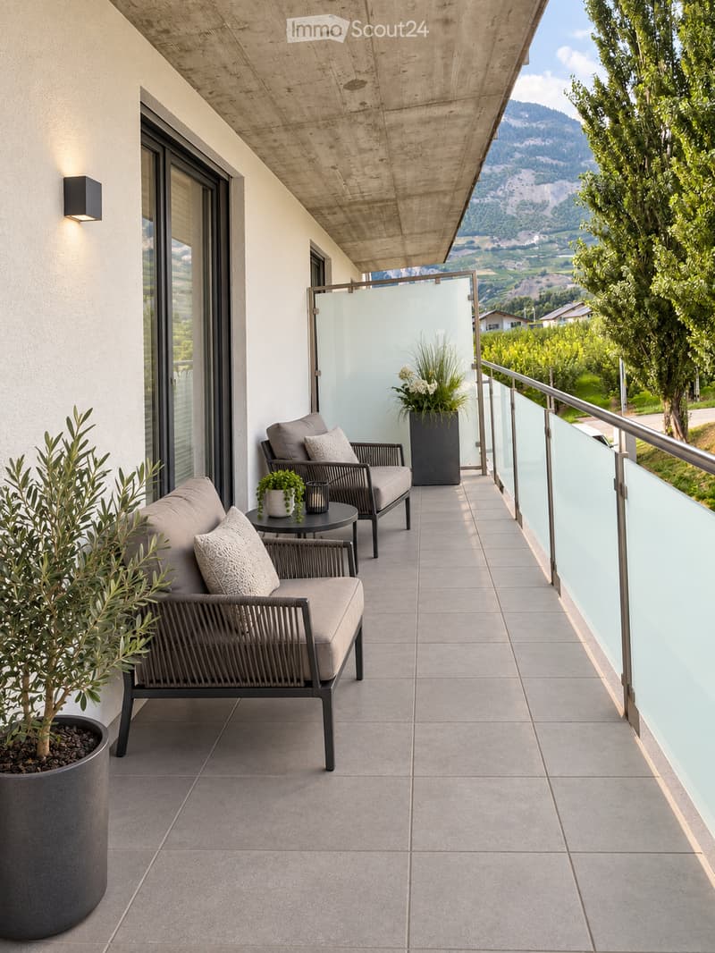 Modern balcony with grey wicker sofa, matching chair, small round table, and potted plants, overlooking a scenic valley and mountains.