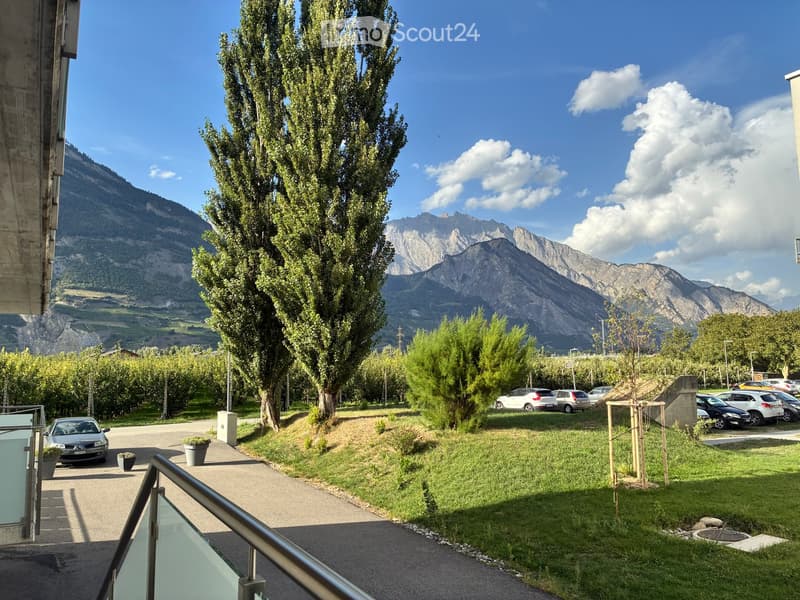 View from a balcony showing a parking area with several cars, a grassy lawn, trees, and a scenic mountain backdrop under a partly cloudy sky.