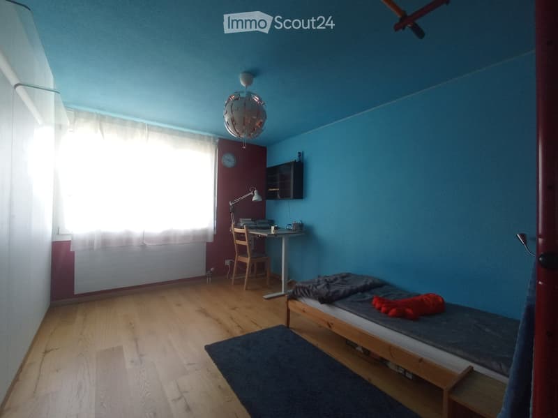 Cozy bedroom with wooden floor, blue walls, a single bed with a dark blue duvet and red pillow, a small desk with a chair, a wall-mounted lamp, and a window with white curtains.