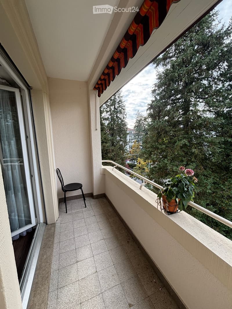 Balcony with tiled floor, white railing, and a view of greenery. Features a black chair and a flower pot with pink flowers on the railing.