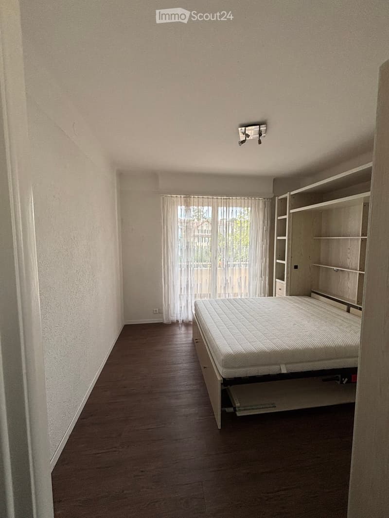 Minimalist bedroom with a single bed, built-in shelving unit, and large window with sheer curtains.