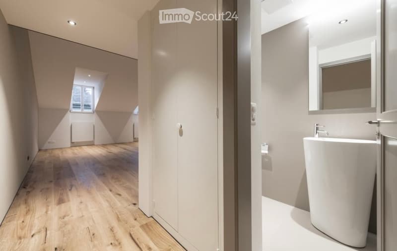 Bright, minimalist hallway with wooden flooring and white walls, leading to a modern bathroom featuring a sleek sink and mirror.