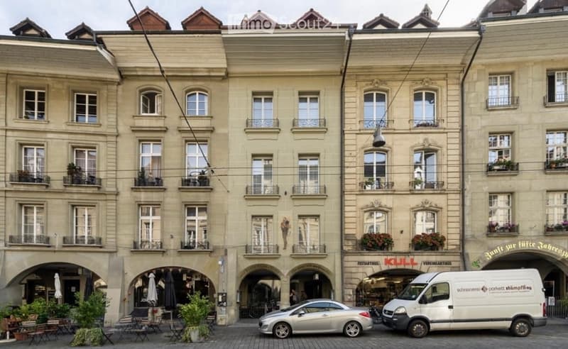 Three-story building with classic European architecture, multiple windows, balconies, and storefronts. A car and van are parked in front.