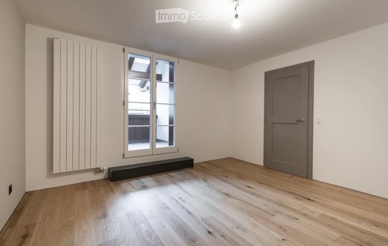 Empty room with wooden flooring, white walls, a grey door, and a large window with glass panels. A radiator is visible on the left wall.