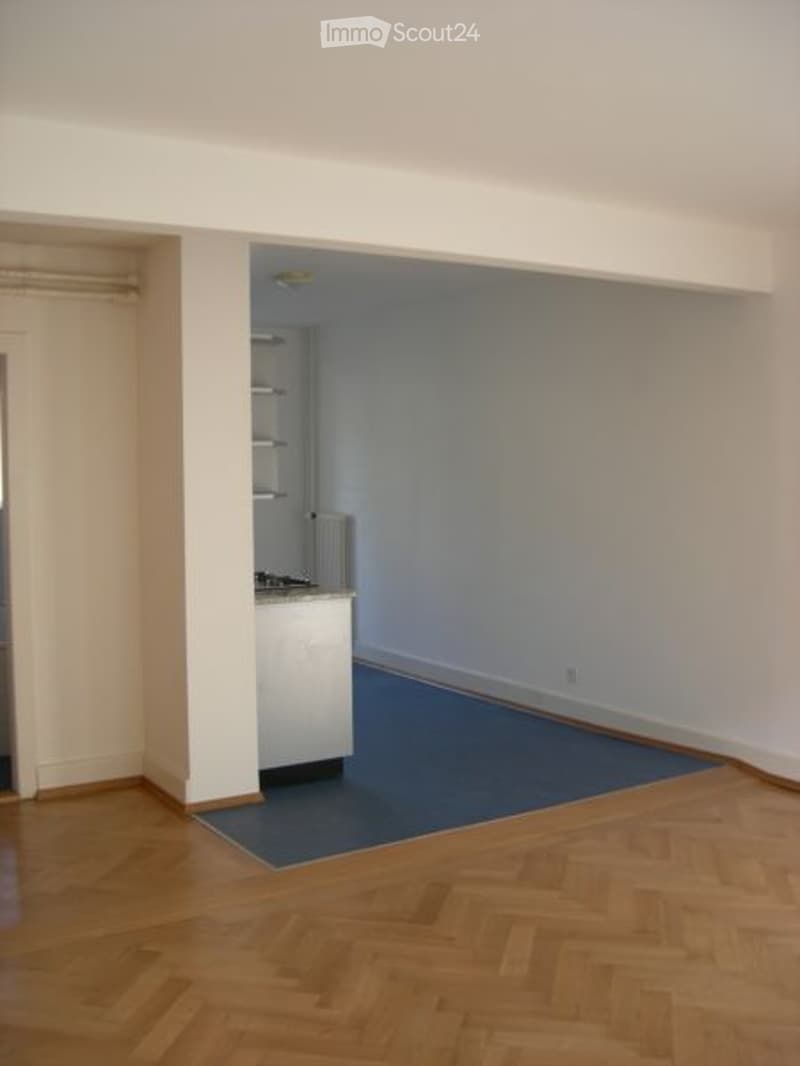 Empty room with wooden flooring, white walls, and a small kitchen area in the corner.