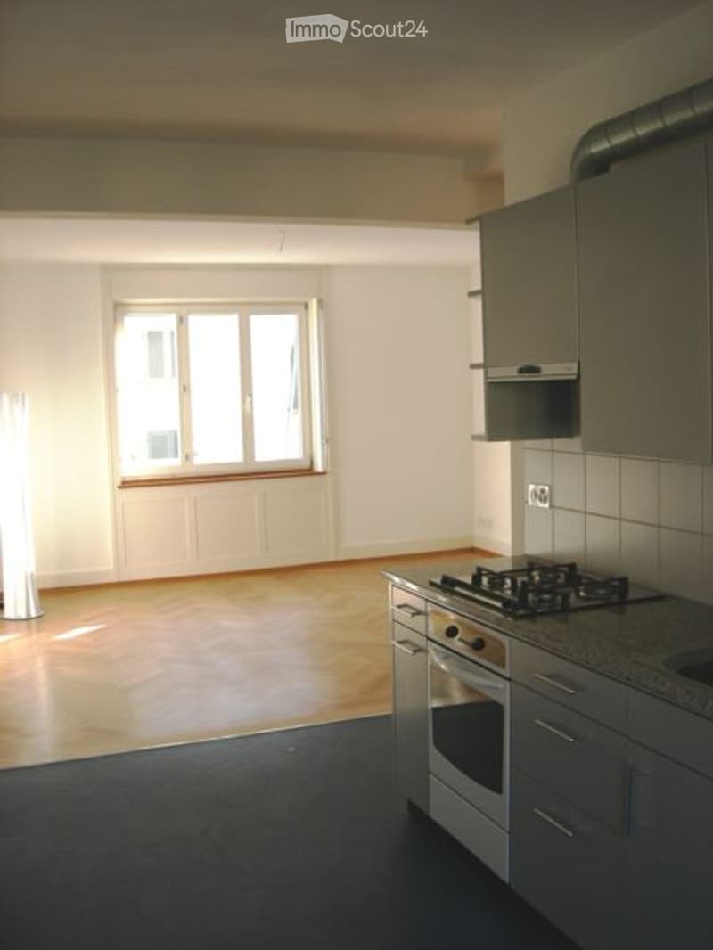 Empty kitchen with wooden floor, white walls, and a window. No furniture or appliances are present.
