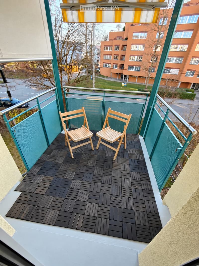 Balcony with dark wooden decking, two wooden chairs, green metal railing, and a view of a residential area with buildings and trees.