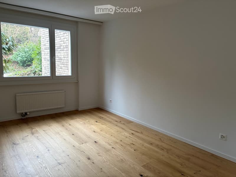 Empty room with wooden flooring, white walls, and large windows letting in natural light. A radiator is visible on one wall.