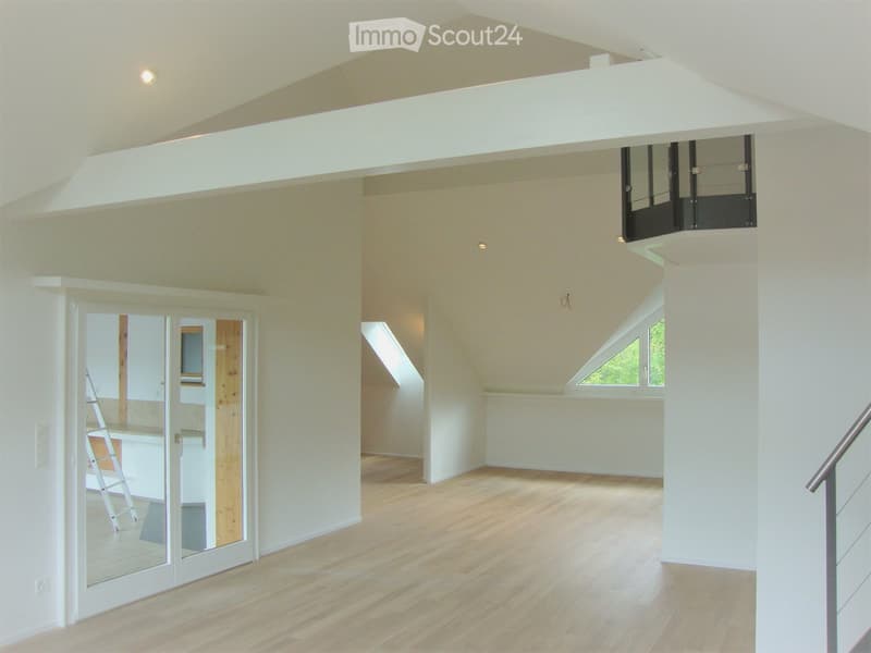 Bright, open loft space with wooden flooring, white walls, and large windows. Features a modern, raised ceiling design and a visible staircase leading to an upper level.