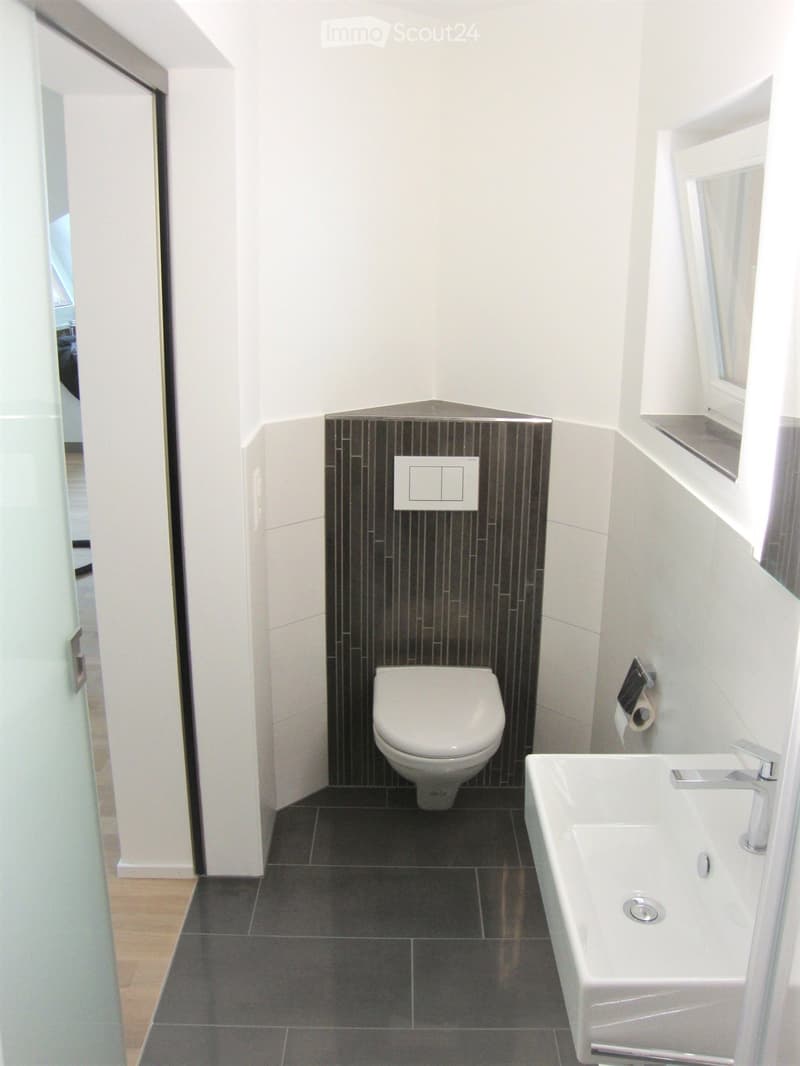 Modern bathroom with white walls, dark tiled floor, toilet, sink, and a window with a shelf above it.