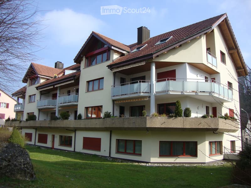 Two-story house with beige exterior, red shutters, multiple balconies, and a well-maintained lawn.