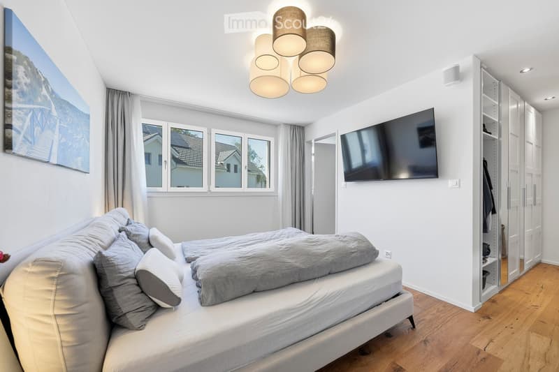 Modern bedroom with a neatly made bed, built-in wardrobe, wall-mounted TV, large windows with curtains, and a scenic picture on the wall.