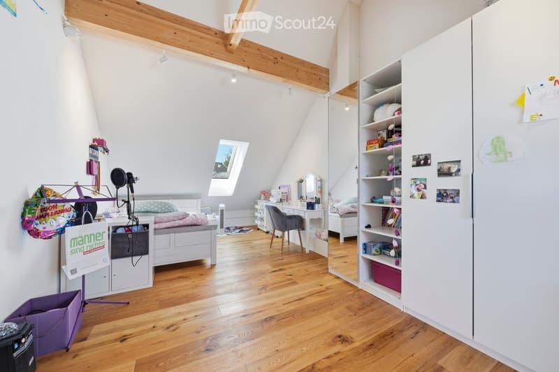 Bright attic bedroom with wooden floors, slanted ceilings, and large skylight. Features a white bed with storage drawers, a white shelving unit, a small desk with a chair, and a vanity area with a mirror.