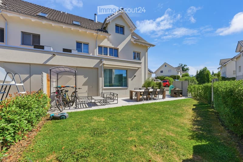 Well-maintained garden with a lawn, patio area with a dining table and chairs, bicycles, and a small shed. The house in the background is a two-story structure with a gray roof and white walls.