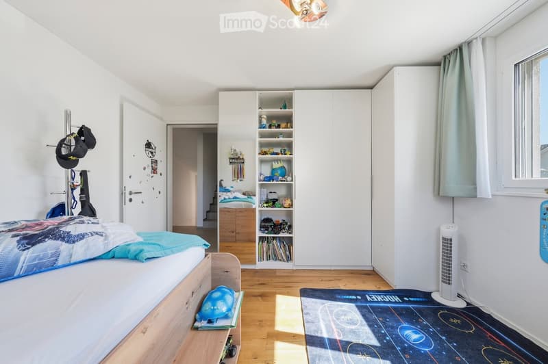 Bright bedroom with wooden flooring, white walls, a bed with colorful bedding, a large wardrobe with shelves, a fan, and a rug with a space-themed design.