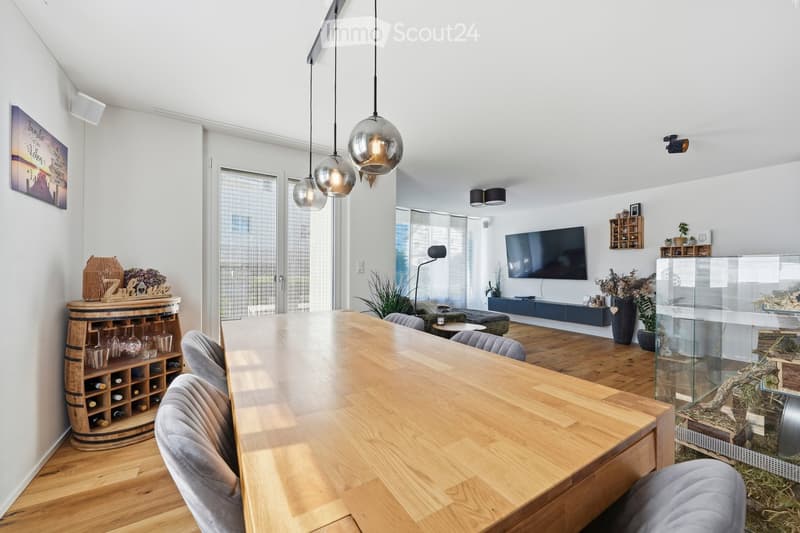 Modern living and dining area with a long wooden dining table, grey upholstered chairs, a wine barrel wine rack, a flat-screen TV, floor lamp, and decorative plants.