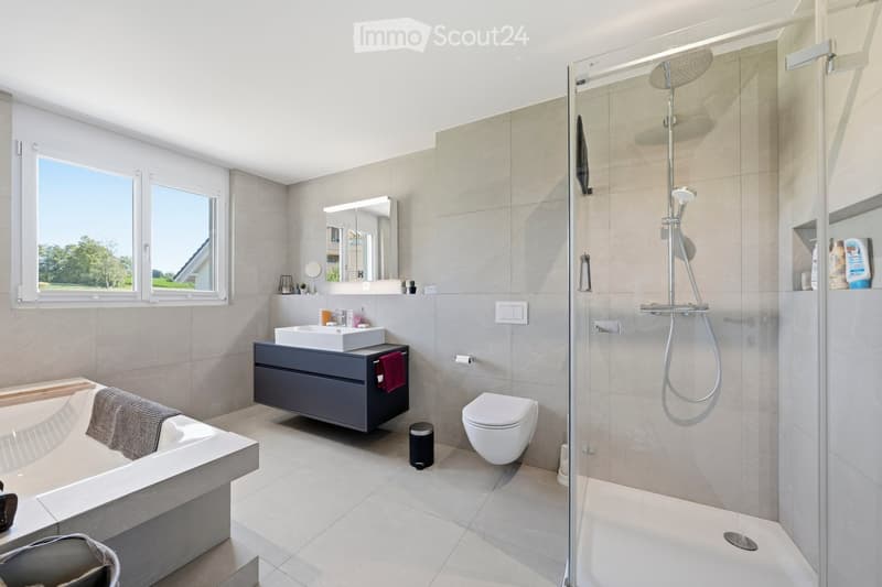 Modern bathroom with a bathtub, sink, toilet, and walk-in shower. Features large windows, grey tiles, and a sleek vanity with a mirror.