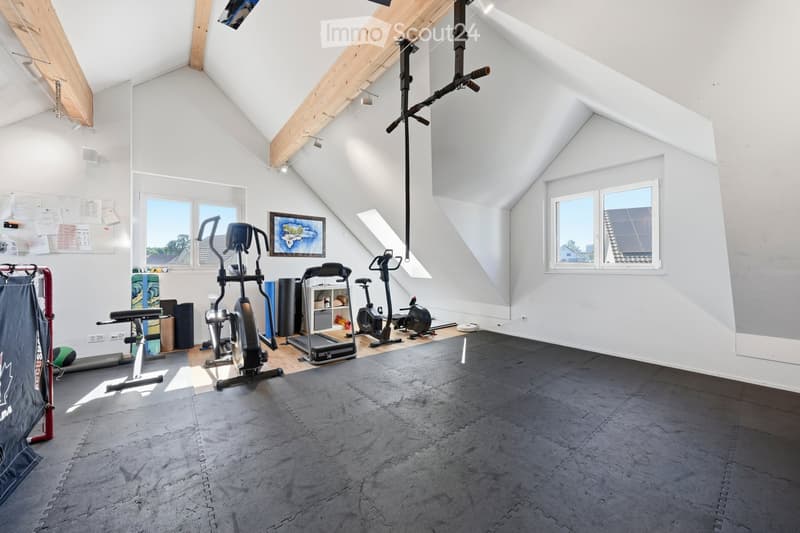 Well-lit attic gym with wooden beams, large windows, and various exercise equipment including a treadmill, elliptical machine, exercise bike, weight machine, and a ball. The space features a grey rubber flooring and a small shelf with items.