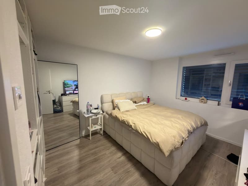 Well-lit bedroom with a large bed, nightstand, mirror, and television. Wooden flooring and white walls.