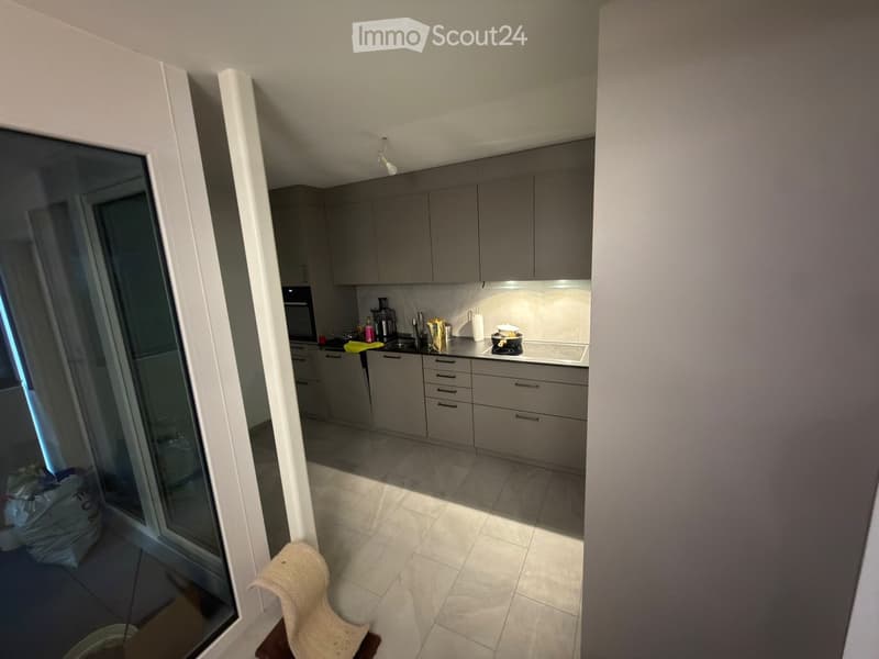 Modern kitchen with grey cabinets, a small chair, and a stool near the entrance. The kitchen features a sink, oven, and various small appliances on the counter.
