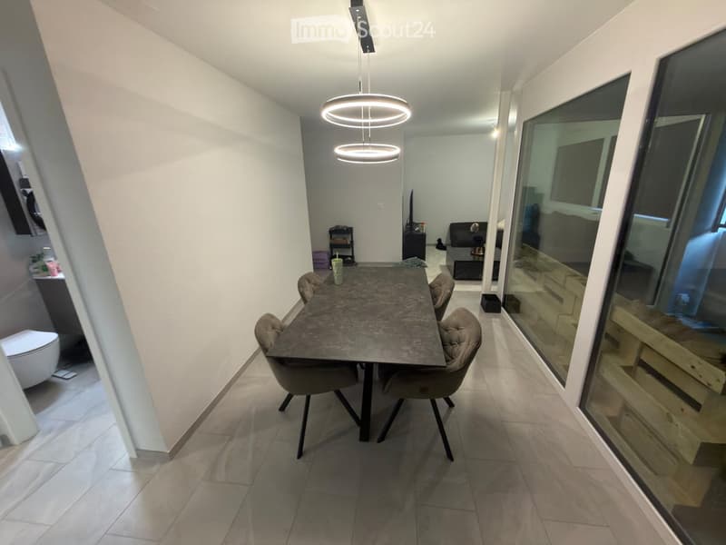 Modern dining area with a dark marble dining table, four chairs, and a hanging circular light fixture. The space is connected to a living area with a sofa and a small shelf.