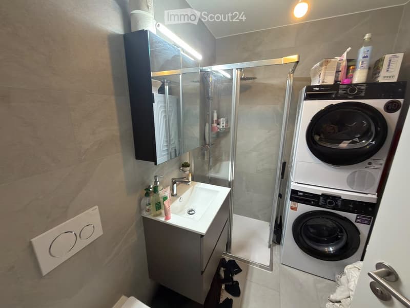 Modern bathroom with a sink, mirror, shower, and a stacked washing machine and dryer. The sink has various toiletries, and the shower area is enclosed with a glass door.