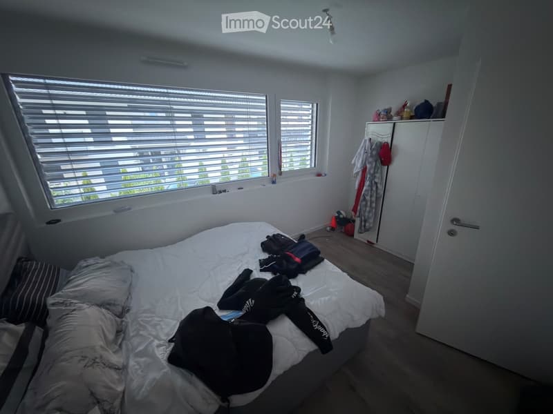 Bedroom with a bed, pillows, a wardrobe with a mirror, and some clothing items on the bed and floor.
