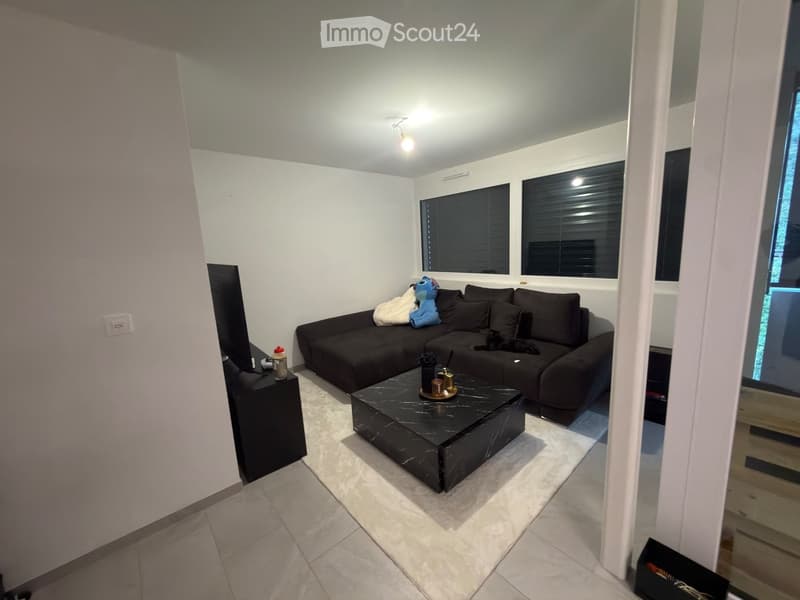 Modern living room with a black sectional sofa, a black coffee table, a TV stand, and a small table lamp. The room features large windows with blinds and a tiled floor.