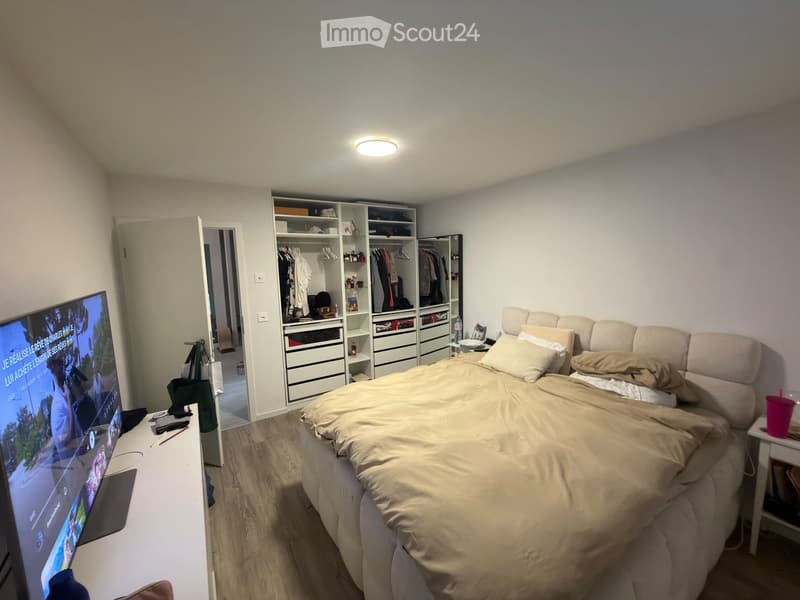 A modern bedroom with a large bed, beige bedding, a nightstand, a television, a wardrobe, and a laptop on a desk.