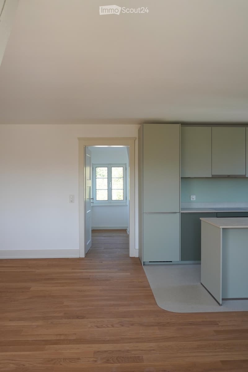 Empty room with wooden flooring, white walls, and a doorway leading to another room with a window. Kitchen area visible with cabinets and a countertop.