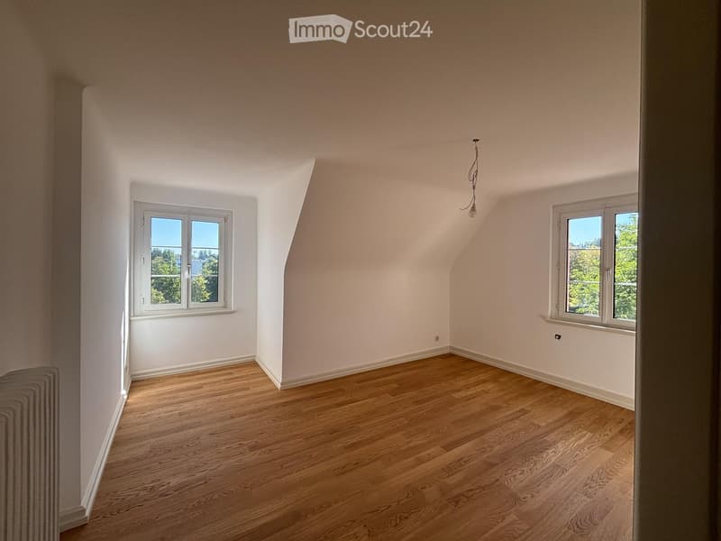 Empty attic room with wooden flooring, white walls, and two windows offering a view of greenery outside.