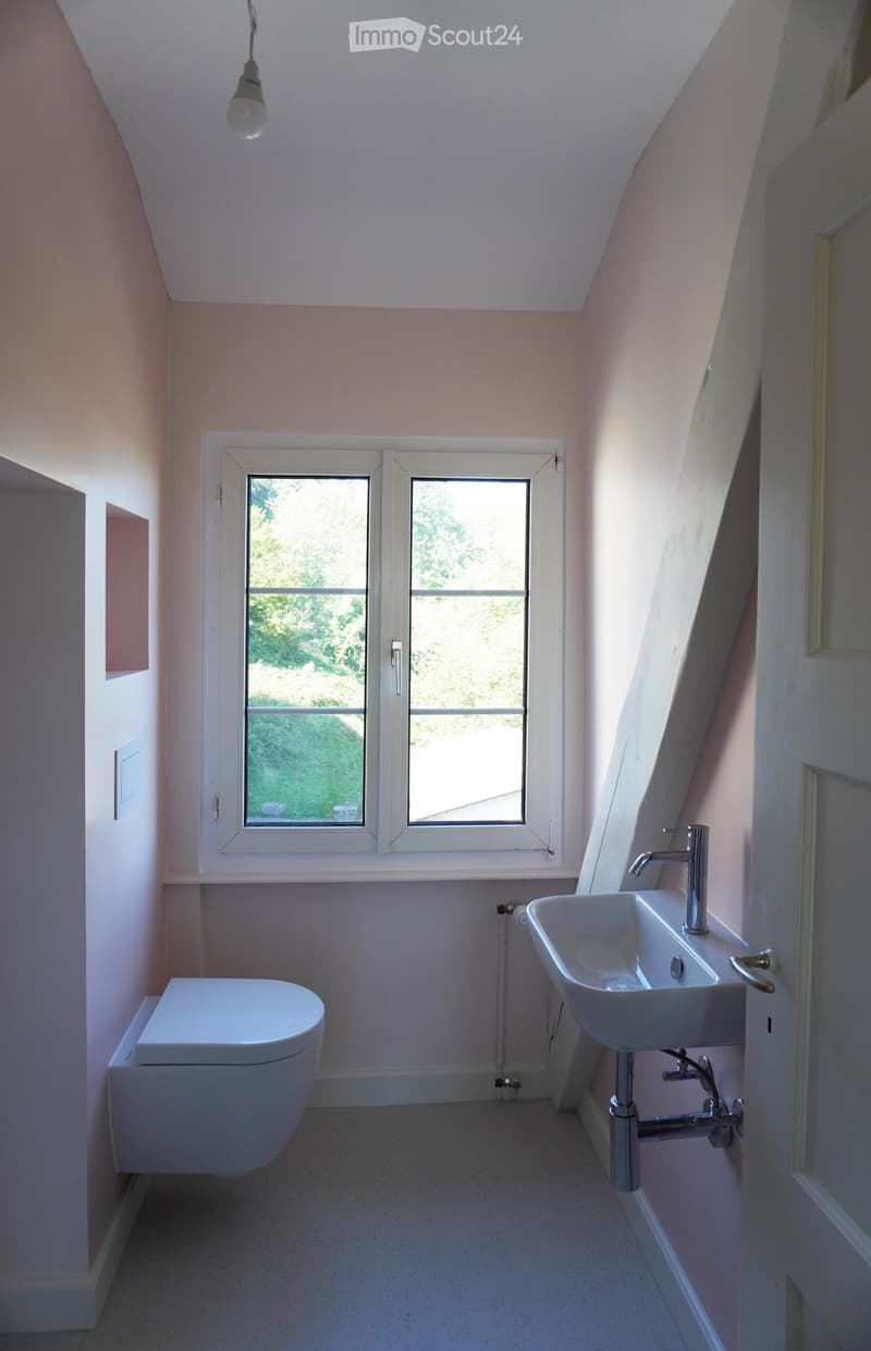 Modern bathroom with a toilet, sink, and window, featuring light-colored walls and a sleek design.