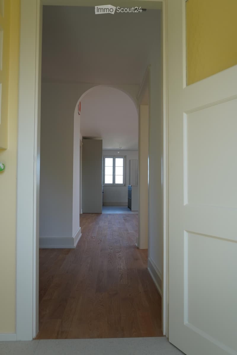 Empty hallway with wooden flooring, white walls, and an arched doorway leading to another room with a window and radiator.