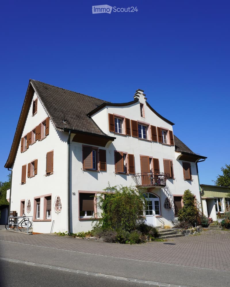 Two-story house with white walls, brown shutters, and a tiled roof. Features include a balcony on the upper floor, a small garden area, and a paved driveway.