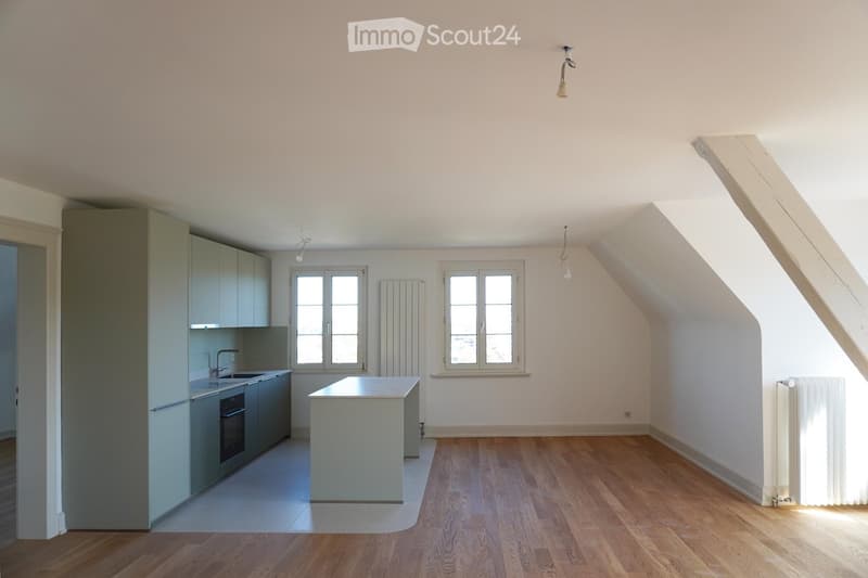 An unfurnished room with wooden flooring, white walls, and a small kitchen area featuring cabinets, a sink, and a countertop. The room has large windows and a slanted ceiling typical of an attic space.