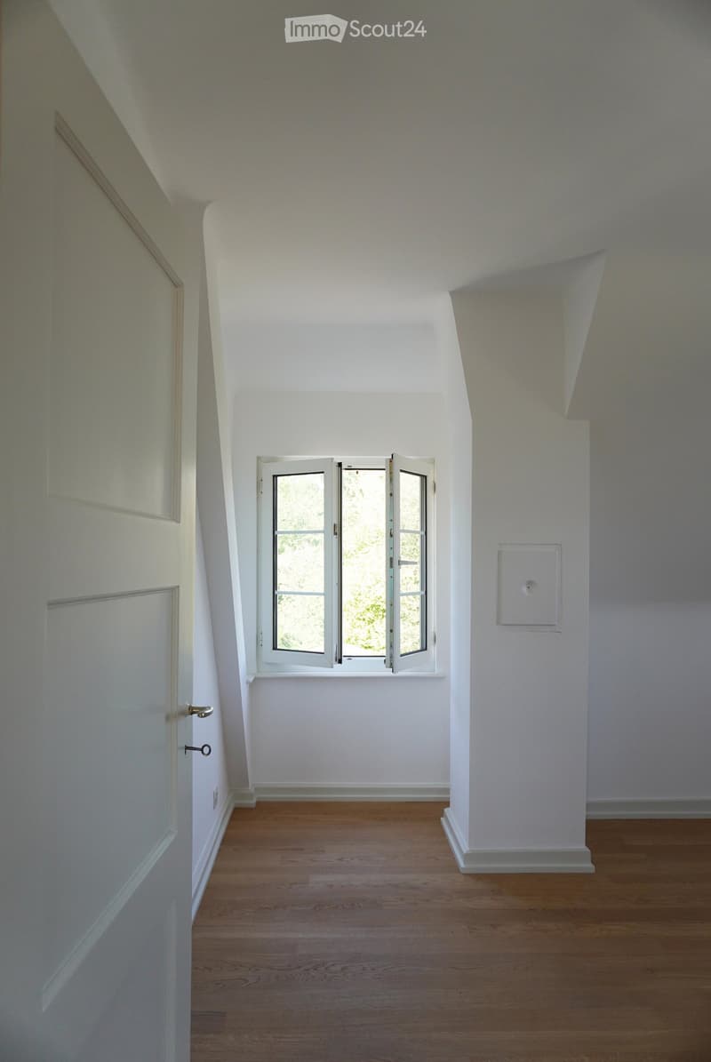Empty room with wooden flooring, white walls, and a window with white frames. The ceiling has a sloped design typical of an attic space.