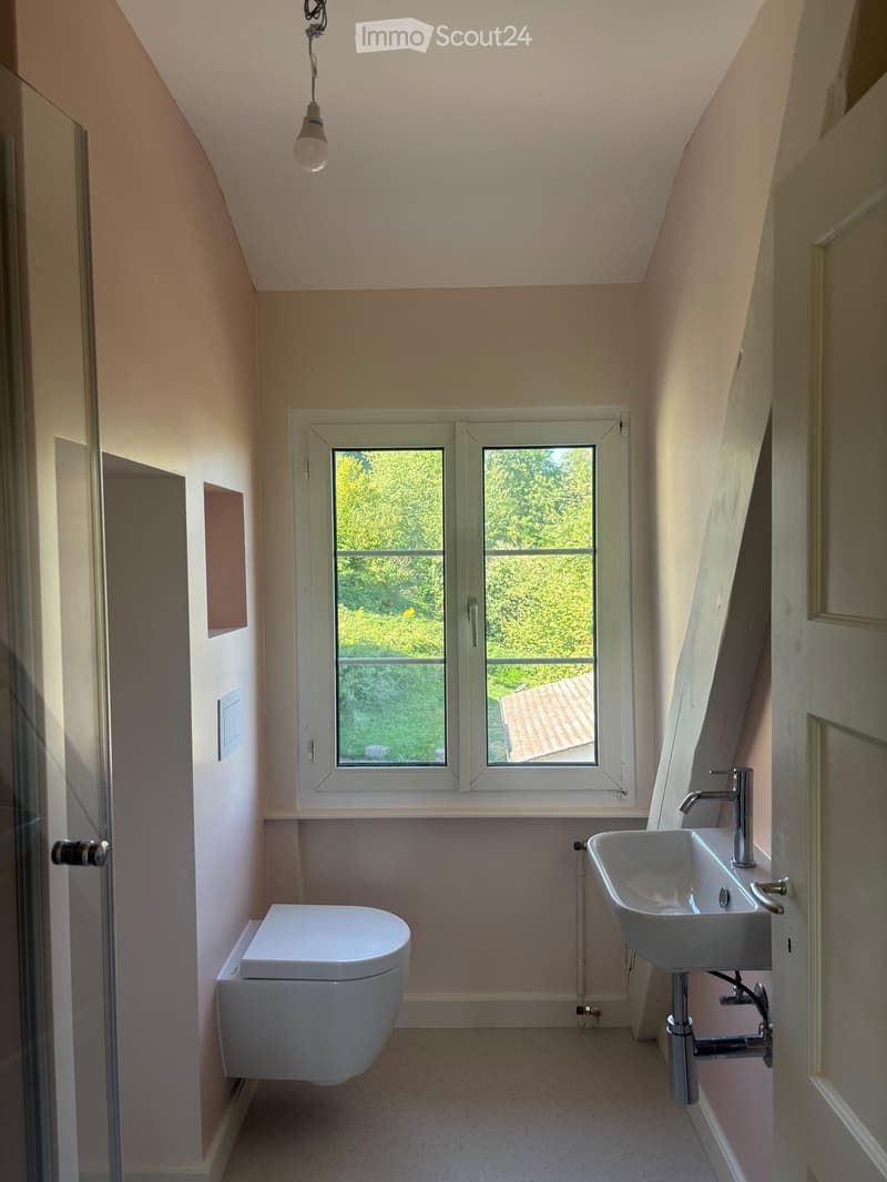 Modern bathroom with a toilet, sink, and window overlooking greenery. The room features light-colored walls and a sleek design.