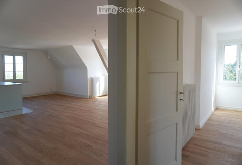 Empty room with wooden flooring, white walls, and large windows allowing natural light. The room features a slanted ceiling typical of an attic space, with radiators visible along the walls.