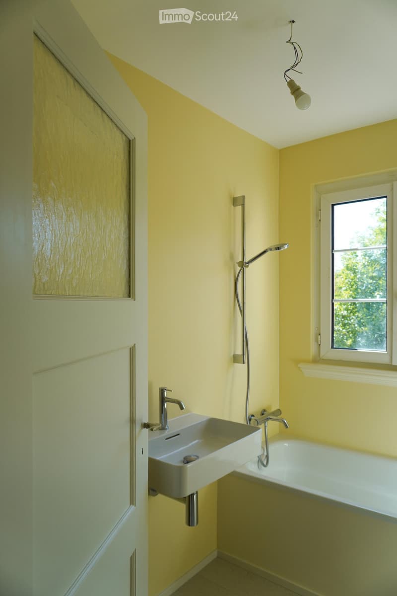 Bright yellow-painted bathroom with a white sink, modern faucet, bathtub, and a showerhead mounted on the wall. A window provides natural light, and a door is visible on the left side.