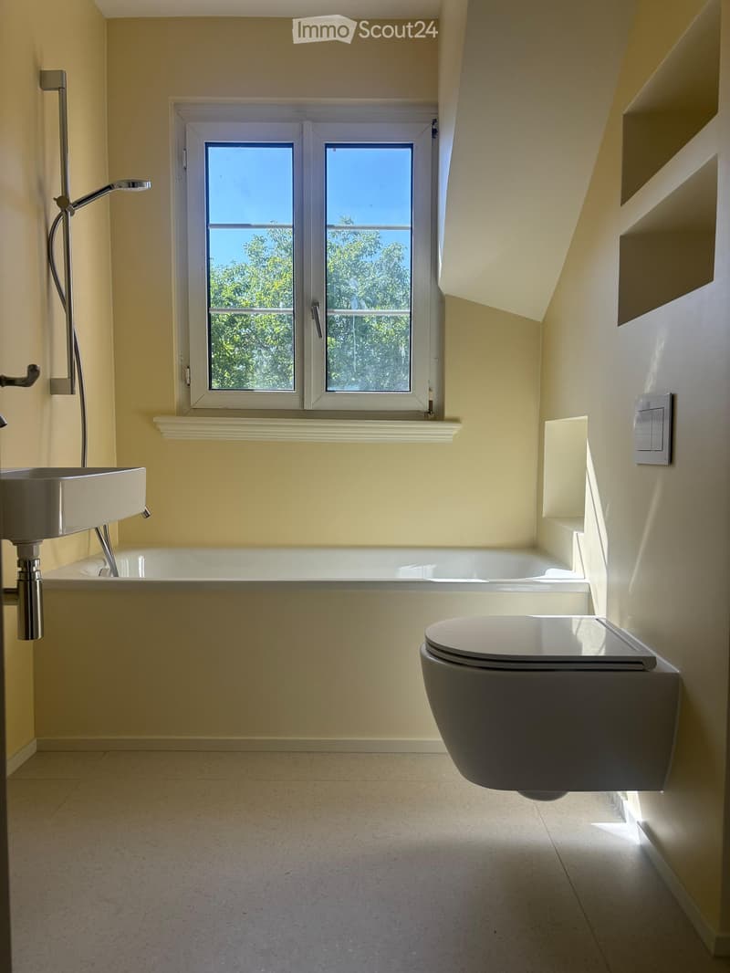 Modern bathroom with a bathtub, toilet, and sink, featuring a large window with a view of greenery outside.