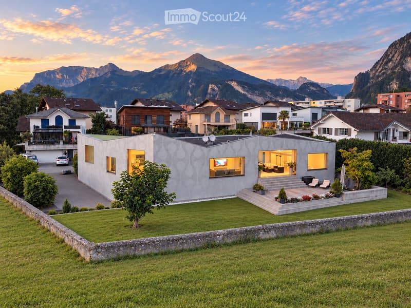 Modern single-story house with a textured facade, large windows, and a well-maintained lawn in a scenic mountain setting.
