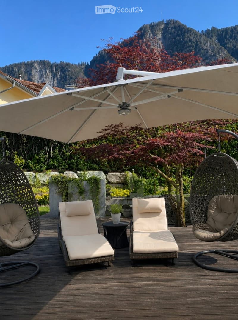 A wooden deck with lounge chairs, hanging egg chairs, a large umbrella, and a small table with a potted plant, set against a backdrop of mountains and greenery.