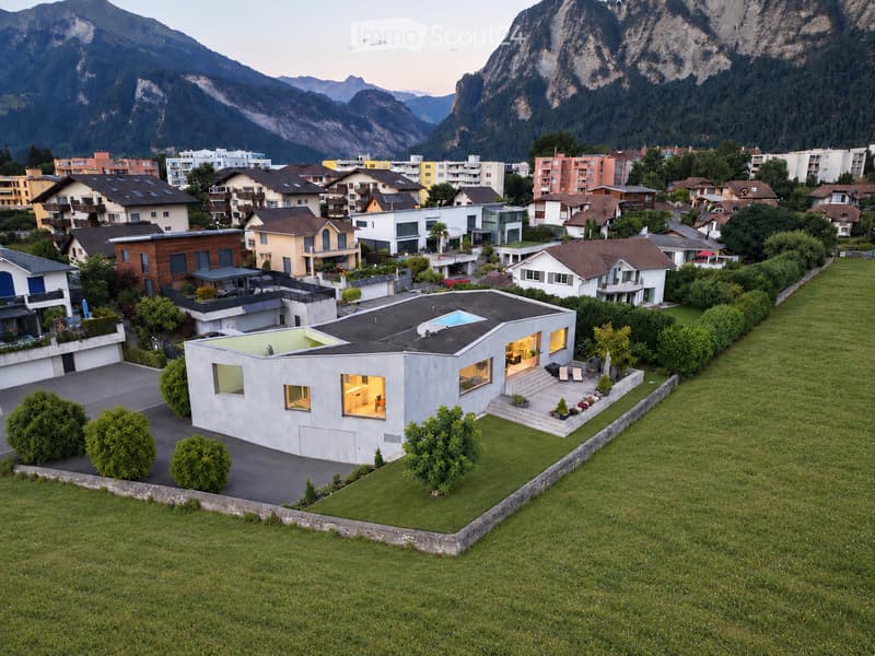 Modern single-family house with a flat roof, large windows, and a small swimming pool on the roof, surrounded by greenery and other residential buildings in a mountainous area.