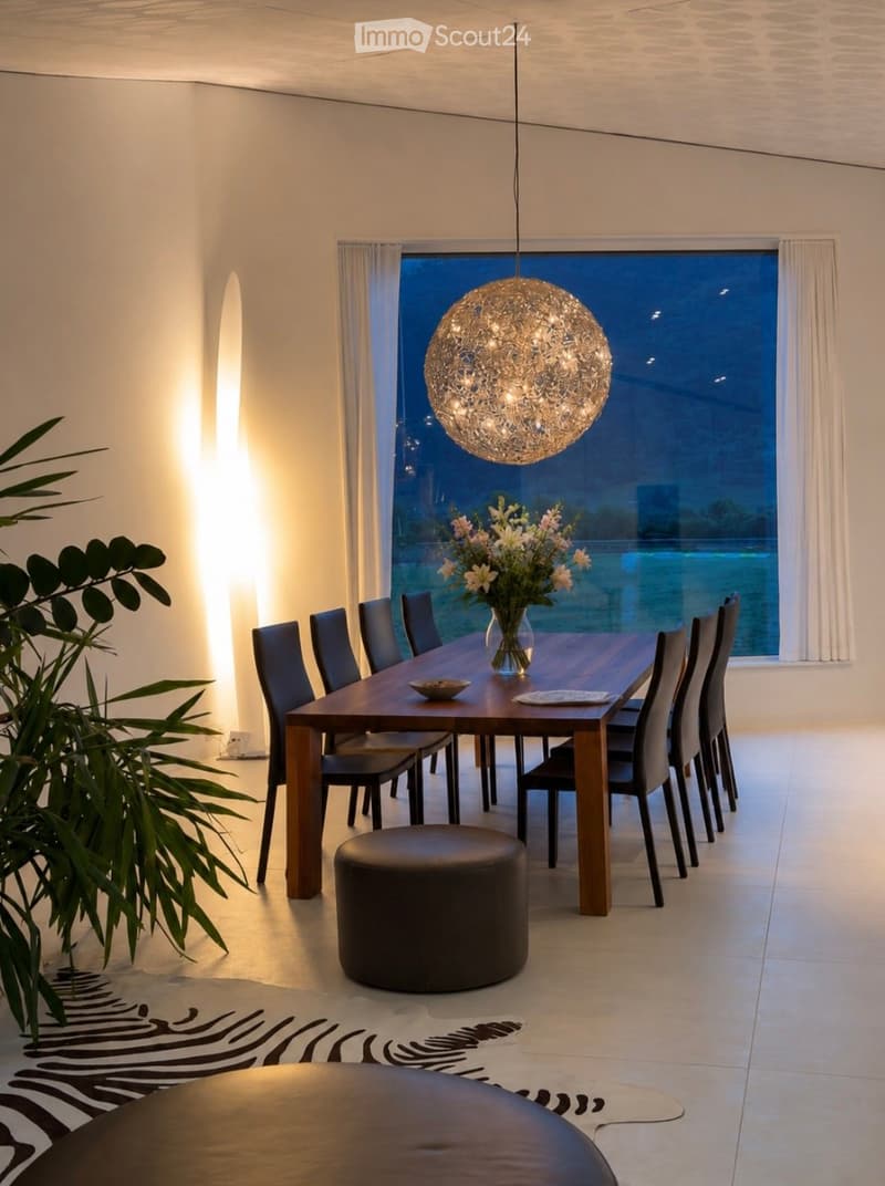 Modern dining room with a long wooden dining table, black chairs, a round black stool, a vase with flowers, and a large spherical pendant light hanging from the ceiling. The room features large windows with white curtains and a view of the night sky.